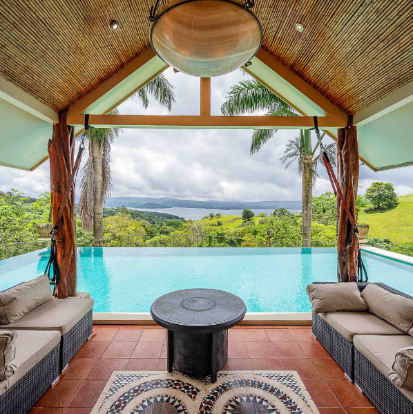 Covered lounge area facing an infinity pool with panoramic views of Lake Arenal, rolling green hills, and tall palm trees.