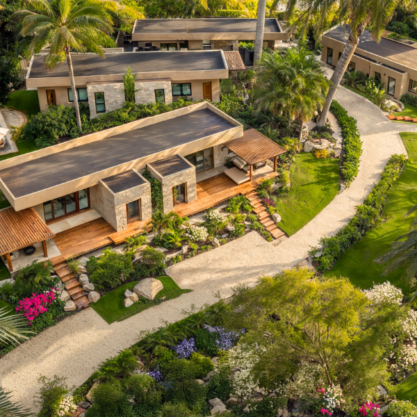 Aerial view of modern tropical villas with flat roofs, natural stone and wood finishes, surrounded by lush landscaped gardens, palm trees, and winding gravel pathways in a serene community setting.