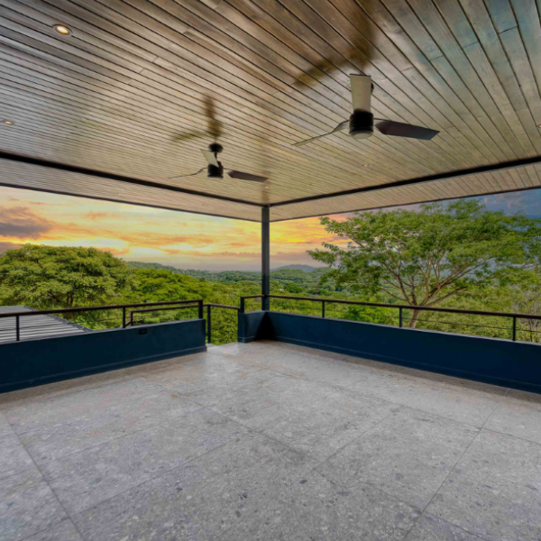 Covered terrace with a wood-paneled ceiling and ceiling fans, opening to expansive sunset views over rolling green hills and treetops in a tranquil natural setting.