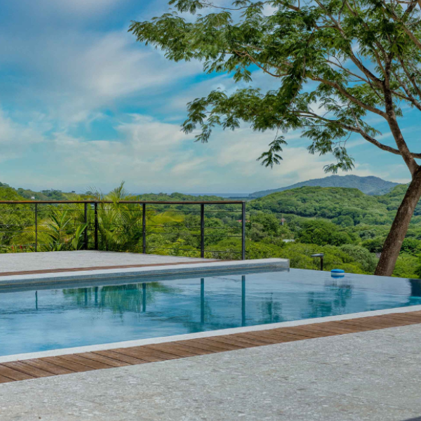 A modern outdoor pool terrace overlooking lush green hills under a bright blue sky, with tropical trees and a sleek black railing framing the natural landscape.