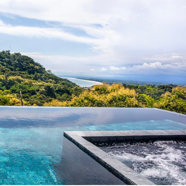 An infinity-edge pool and jacuzzi overlooking lush rainforest and a distant ocean coastline under a bright sky.