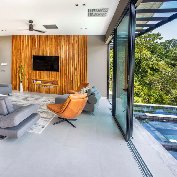 A contemporary living room with full-height sliding glass doors, a wooden feature wall with a mounted TV, and views of the jungle canopy.