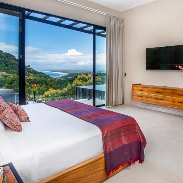 A bedroom with floor-to-ceiling glass doors opening to ocean and rainforest views, featuring a minimalist interior with wood furniture.