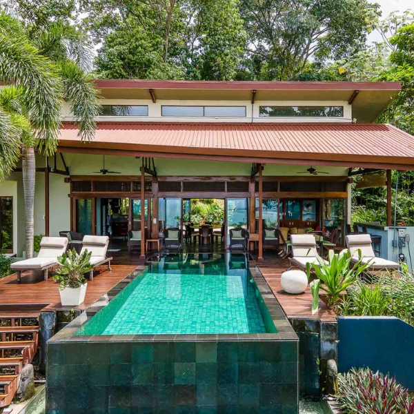 Modern house with infinity pool in Uvita, wooden deck, sun loungers, open design, surrounded by lush tropical gardens and palm trees.