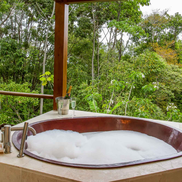 Outdoor jacuzzi tub filled with bubbles, champagne and glasses on the side, overlooking dense tropical forest from private terrace.