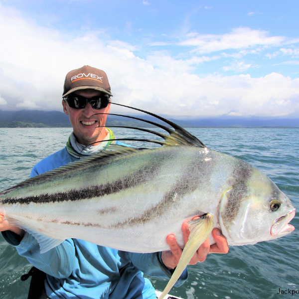 A man holding a big roosterfish with the ocean and coastline in the background.