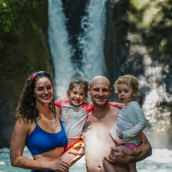 alt: A family of four smiles in front of a waterfall, with two adults holding two young children in shallow water.