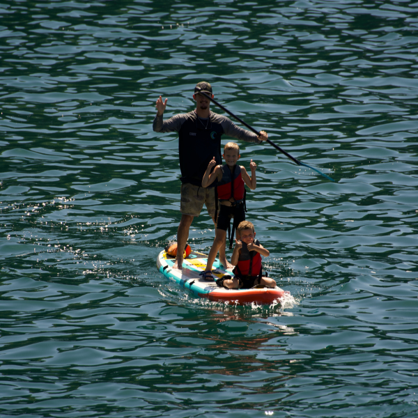alt: An adult and two children ride a stand-up paddleboard together on calm ocean water, all wearing life jackets.