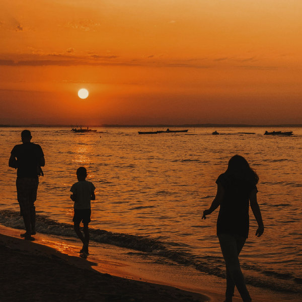 alt: Three people walk along the beach at sunset, silhouetted against an orange sky and calm water with small boats in the distance.