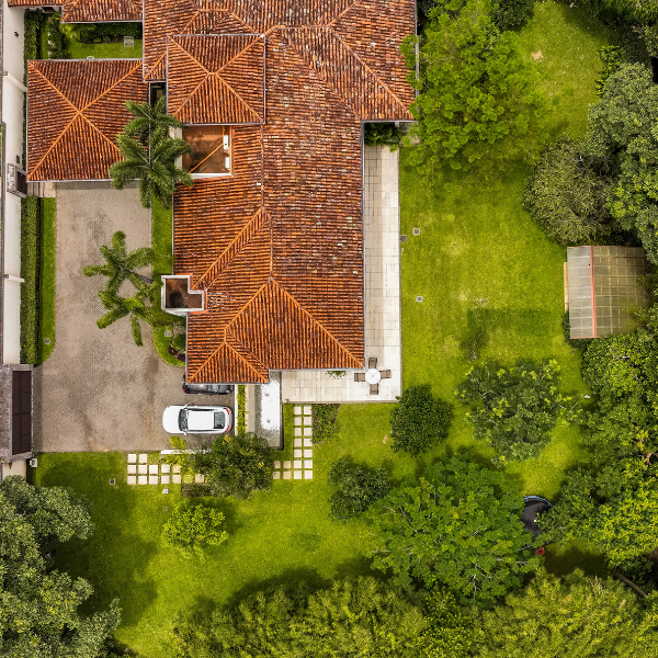 alt: An aerial view of a large home with a red tile roof, long driveway, parked car, and expansive green yard surrounded by trees.