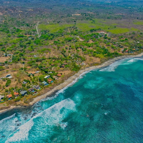 An aerial view of a tropical coastline with turquoise ocean water, white waves breaking along the shore, and scattered homes set back among dry forest and greenery.