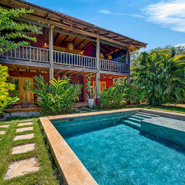 alt: two story tropical style home with a large covered wooden balcony overlooking a rectangular swimming pool, surrounded by lush plants and a sunny garden.