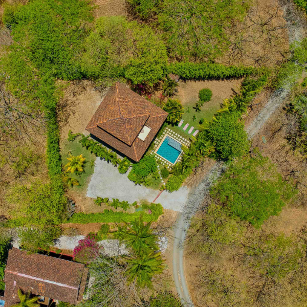 alt: A high aerial view of a private home with a terracotta roof, square pool, landscaped yard, and circular driveway surrounded by dense trees and dry tropical vegetation