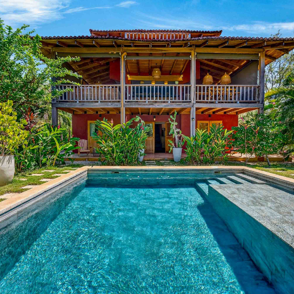 alt: angled exterior view of a tropical two story house with wooden balconies, red toned walls, garden landscaping, and a rectangular pool with wide entry steps in the foreground.