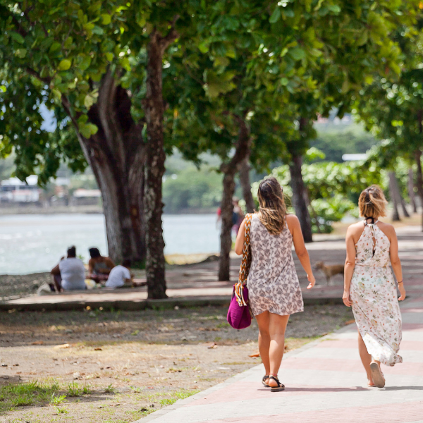 Two women walking along a shaded waterfront path in Costa Rica with trees, locals relaxing, and calm coastal scenery