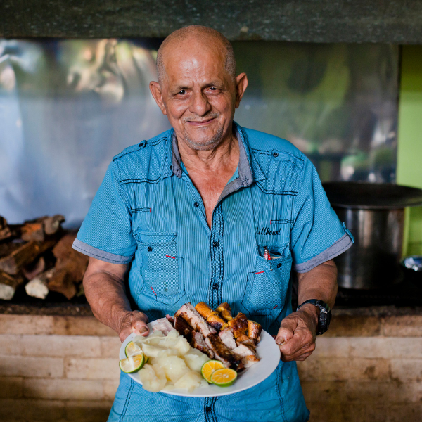 Local Costa Rican man holding a plate of traditional food with grilled meat, yuca, and citrus in a rustic outdoor kitchen
