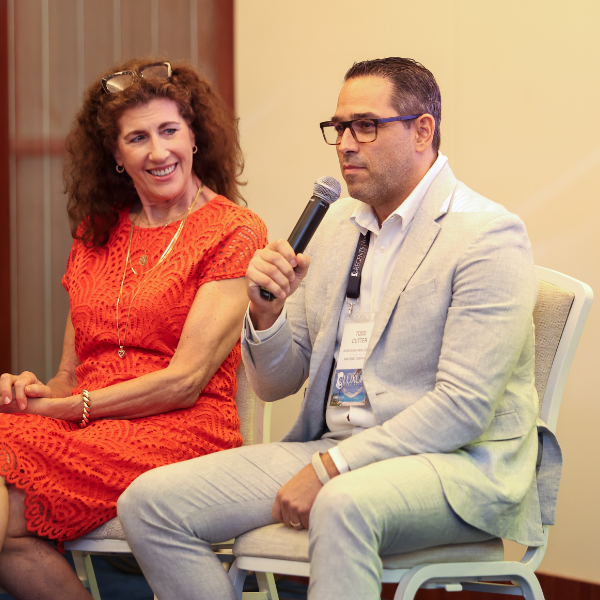 Two people sitting on chairs. The person on the right is holding a microphone and wearing glasses and a light gray blazer. The person on the left is wearing an orange/red dress and smiling.