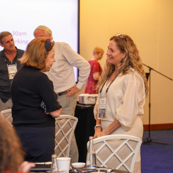 Two women standing and talking to each other indoors. A presentation screen is visible in the background showing partial text. Several other people are visible in the background.