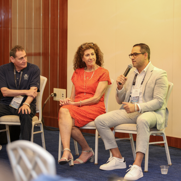 Three people seated on chairs in a row. The person on the right is holding a microphone. Blue carpet is visible on the floor.