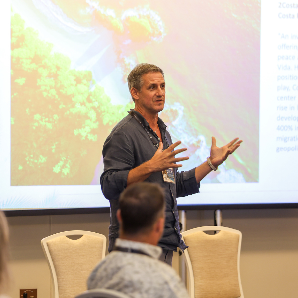 A man standing in front of a large projected presentation slide. The slide shows a colorful aerial/satellite image and text mentioning '2Costa Rica' and 'Costa Rica.' He has both hands raised.