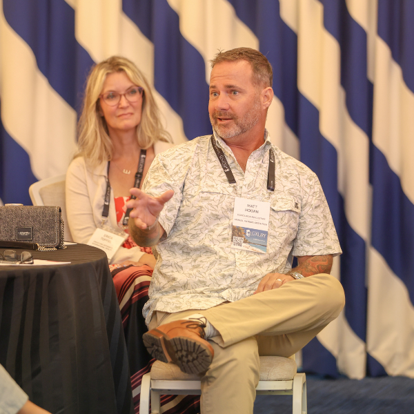 Two people sitting on chairs in front of a blue and white diagonal striped backdrop. The person in the foreground is wearing a patterned short-sleeve shirt and khaki pants, with one hand raised. A laptop bag is visible on a table to the left.