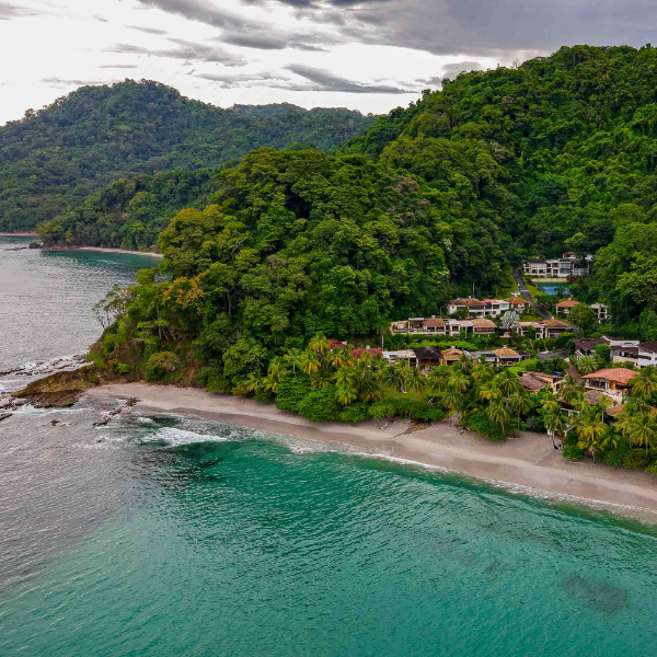 Aerial view of a secluded tropical bay with calm turquoise water, dense green rainforest-covered hills, and a small beachfront residential community tucked along the shoreline.