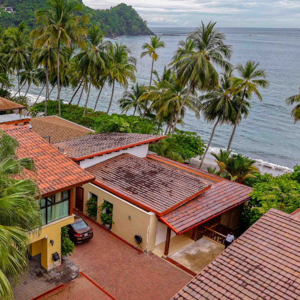 Elevated view of beachfront homes with terracotta tile roofs surrounded by palm trees, directly facing the ocean with gentle waves and a forested headland in the background.