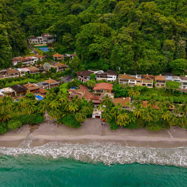 Wide aerial shot of a private beachfront neighborhood featuring multiple luxury residences, palm-lined shoreline, sandy beach, and lush rainforest rising immediately behind the homes.