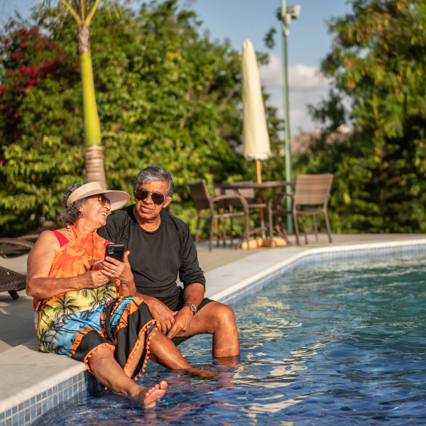 An older couple sitting together at the edge of a swimming pool with their feet in the water, looking at a smartphone and smiling. Lush tropical greenery, a palm tree, and patio furniture are visible in the background.