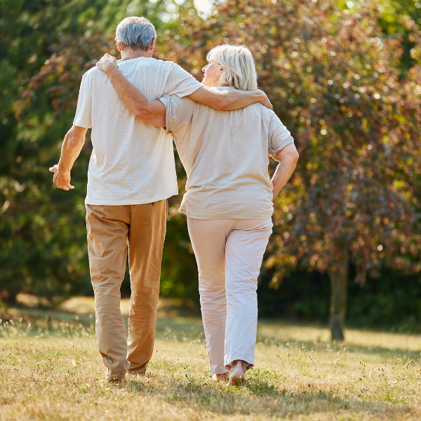 an older couple walking away from the camera through a sunlit park or garden, arms around each other's shoulders. Autumn-toned trees fill the background.