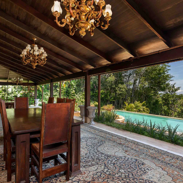 An elegant open air covered terrace or rancho with a dark wood beamed ceiling, two ornate gold chandeliers, and a large dark wood dining table with leather chairs. A decorative mosaic tile floor leads to an open view of a swimming pool and tropical garden beyond.