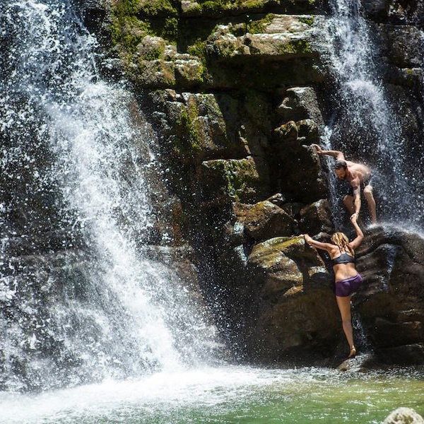 Two people in the middle of a waterfall, one helping the other get on top of a rock.