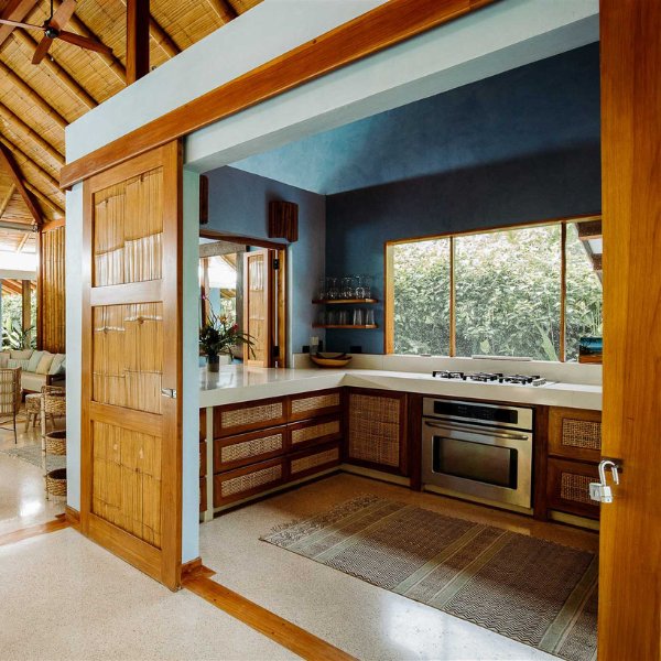 Modern kitchen with blue walls, wooden cabinetry, white countertops, and large window overlooking greenery, designed for tropical luxury living.