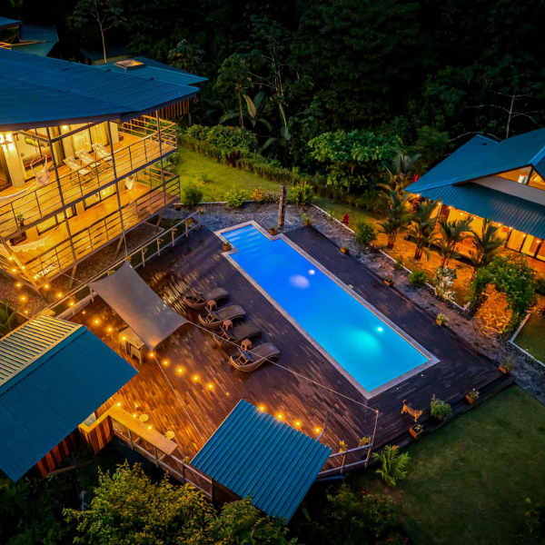 Illuminated infinity pool with lounge chairs and wooden deck at night, framed by modern eco-lodge buildings and lush tropical gardens in Drake Bay, Costa Rica.
