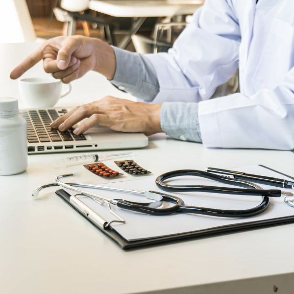 Doctor working at a desk with laptop, stethoscope, prescription medication, and medical clipboard in a clinical office setting.