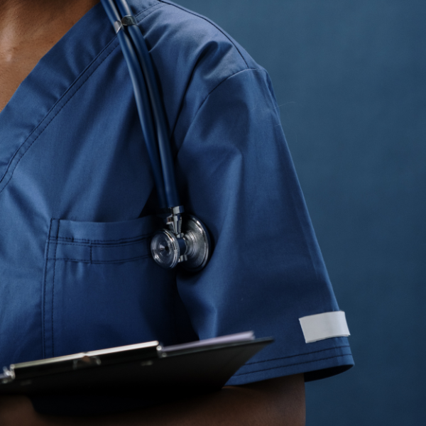 Healthcare professional in blue scrubs holding a clipboard with a stethoscope around their neck against a dark blue background.