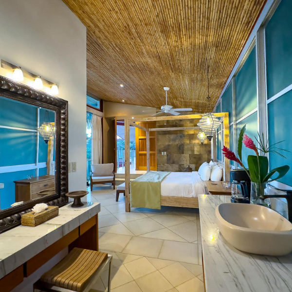 Elegant tropical-style bedroom suite with bamboo ceiling details, four-poster bed, marble vanity, and warm ambient lighting creating a resort inspired atmosphere