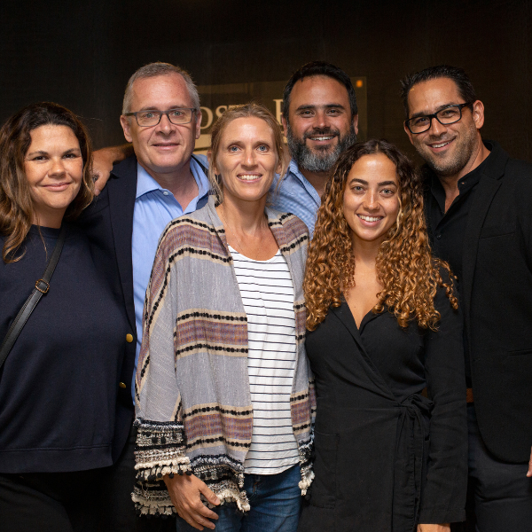 Group portrait of five people smiling together at an indoor networking or company event with dark modern interiors.
