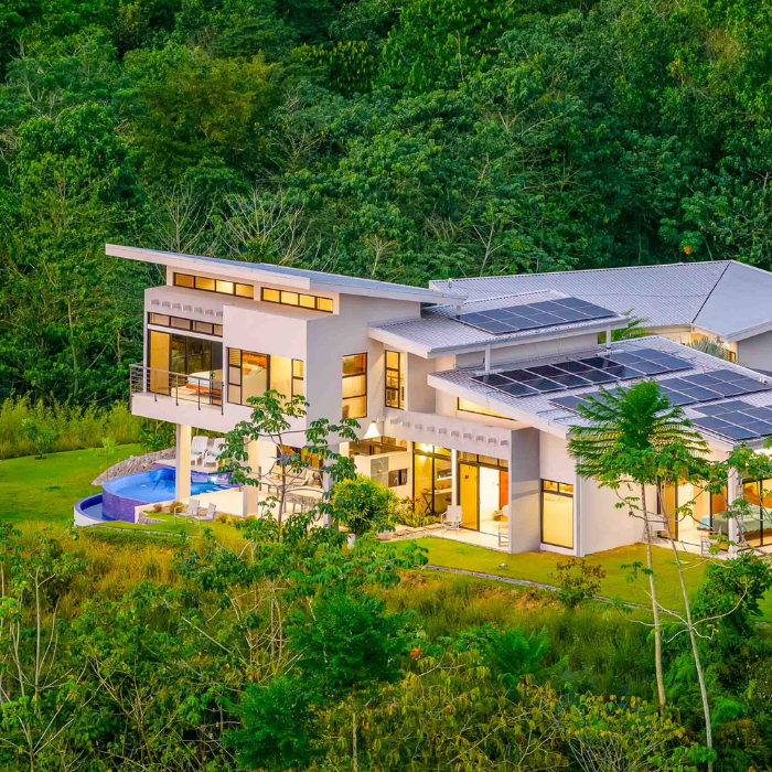 white home with sun panels in the rooftop, surrounded by lush tropical forest, all the lights of the home are turned on and have a potent look