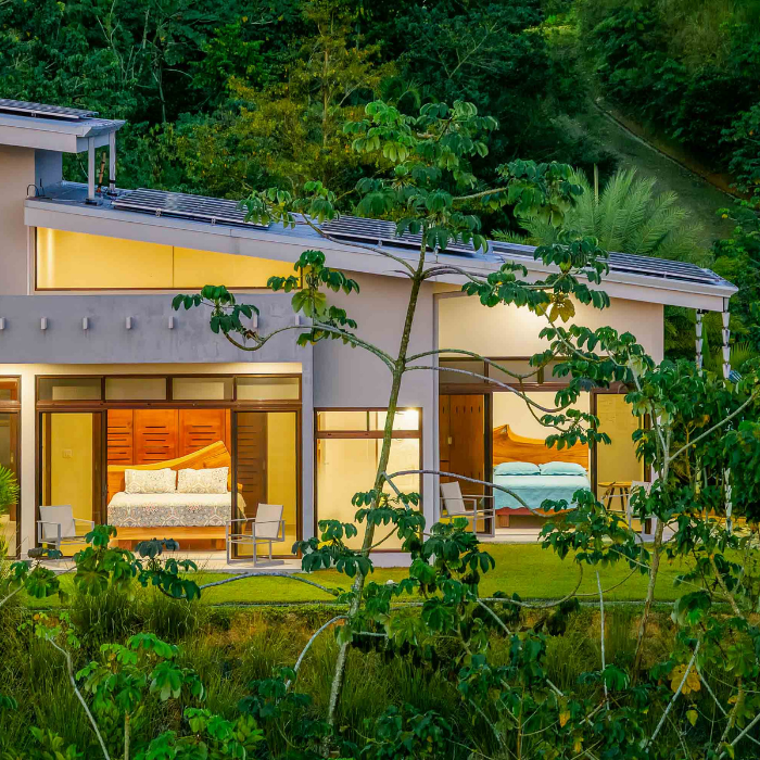 Contemporary guest house bedroom suite with floor-to-ceiling glass doors opening to lush tropical landscaping and private outdoor seating areas.