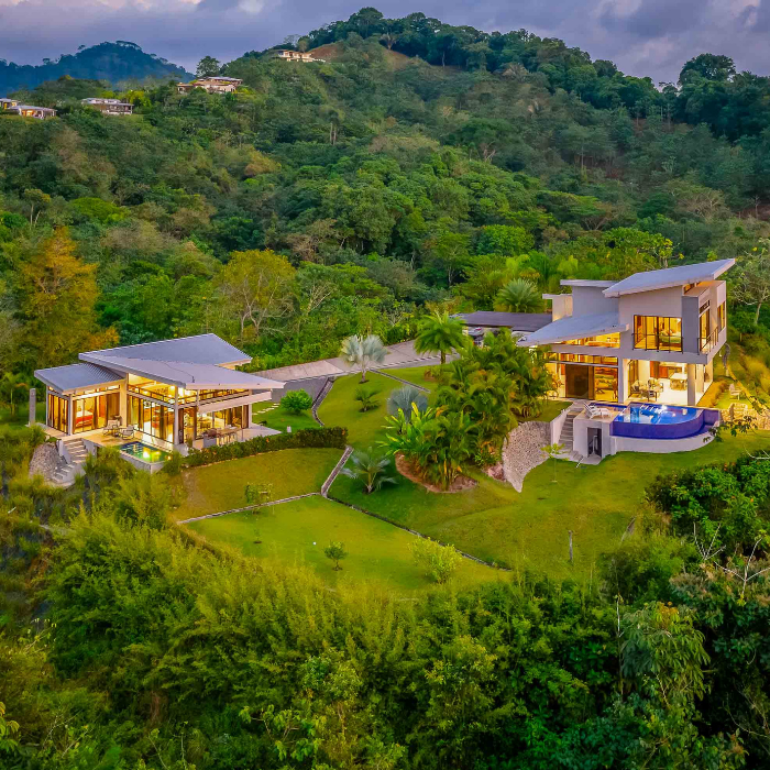 Aerial view of a luxury tropical estate with two modern homes, infinity pool, manicured gardens, and lush rainforest-covered hillsides.