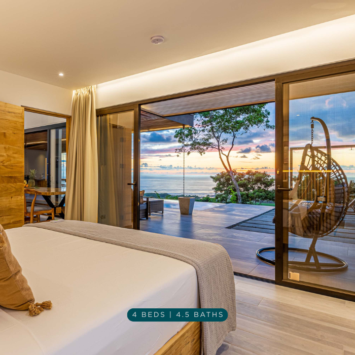 Modern bedroom with floor to ceiling glass doors opening to an ocean view terrace at sunset.